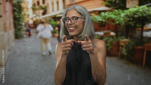 Foto Woman with grey hair points fingers toward camera on a street outside a restaurant terrace, smiling and making playful gesture; joyful warmth