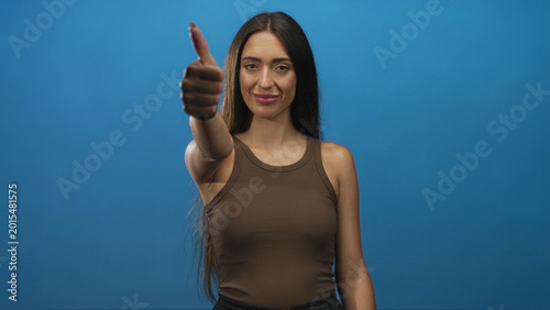 Foto Young hispanic woman shows thumbs up with extended hand and visible thumb in blue studio; approval confidence