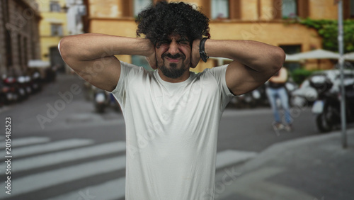 Fotografia Man with raised arms and curly hair standing on a city street crosswalk, looking up with a strained grimace; frustration