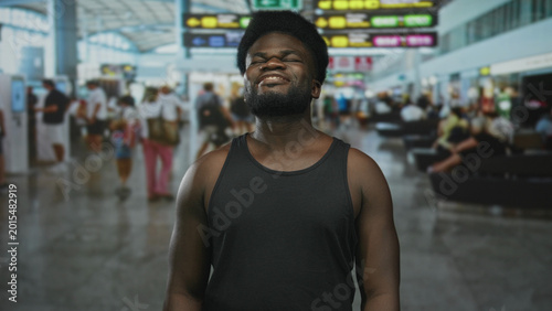 Schilderij op canvas Man with bare shoulders and beard smiling with eyes closed in busy airport terminal wearing black tank top; relief travel ease