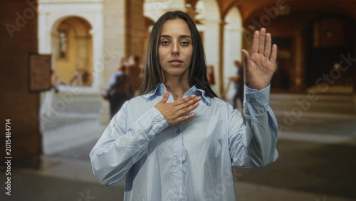Foto Young hispanic woman with hand on chest and right hand raised in old town street building; oath solemnity commitment
