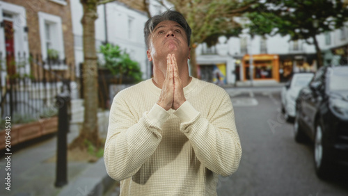 Canvas Print Man with hands pressed together praying on street near parked cars and shopfronts; quiet reflection