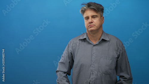 Quadro em tela Man with visible face tilting his head and looking down against blue studio backdrop; melancholy introspection
