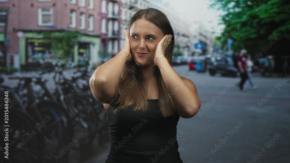 Fototapeta premium Woman covering ears with hands on a city street by parked bicycles, wearing black tank top; urban annoyance.