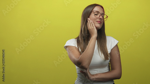 Fotografija Young woman touching cheek with hand and wearing glasses in studio yellow backdrop; thoughtful reflection
