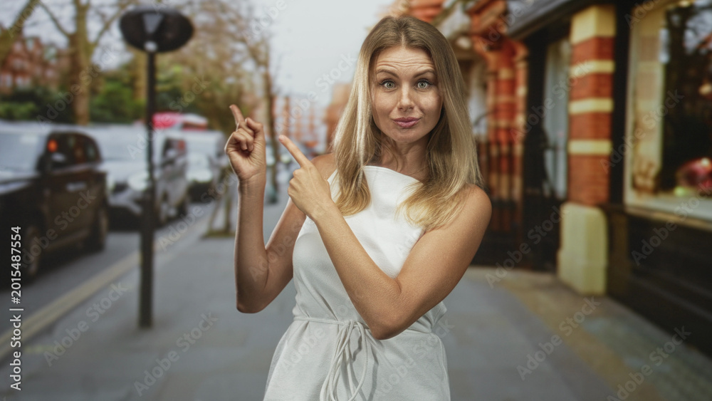 Fototapeta premium Woman points finger to left on city street in front of brick building wearing white dress; playful confidence.