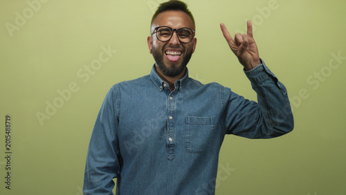Bild auf Leinwand Man with glasses in casual denim shirt flashes rock horns gesture in olive green studio; playful confidence