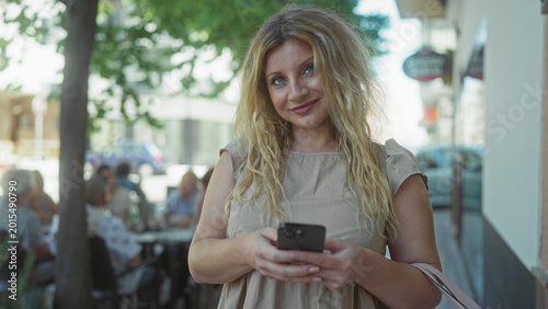 Фотография Woman holding smartphone and smiling on street while checking messages; contentment connection ease