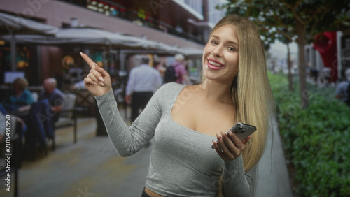 Photos Blonde caucasian woman pointing finger and holding smartphone on street at outdoor cafe, smiling with visible braces; friendly confidence