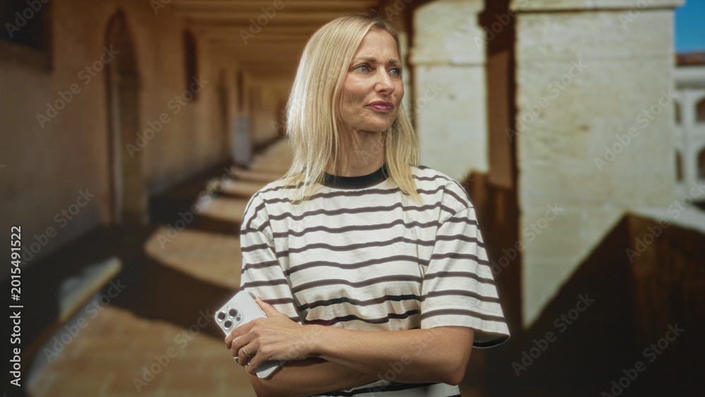Fototapeta premium Woman holding smartphone, arms crossed and looking up in building corridor; contemplation quiet serenity.