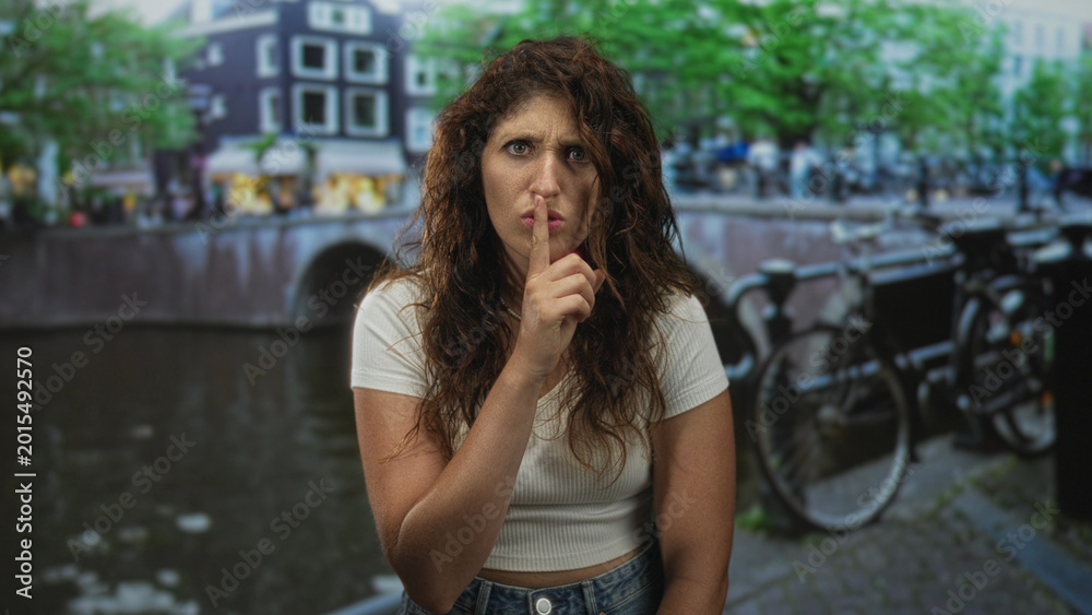 Fototapeta premium Woman with finger to lips for silence on amsterdam street by canal and parked bicycle; quiet secrecy.