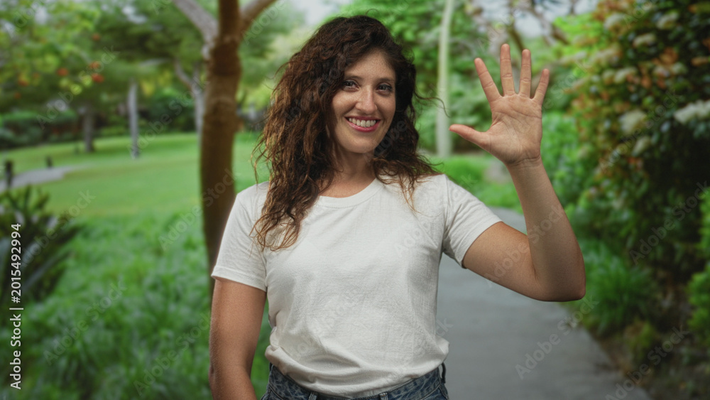 Naklejka premium Woman waving open hand on park path while smiling with palm visible and forearm shown; friendly greeting happy.