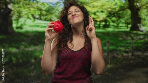Canvastavla Young hispanic woman smiling while holding a red piggy bank and crossing fingers in a green park forest; saving hope
