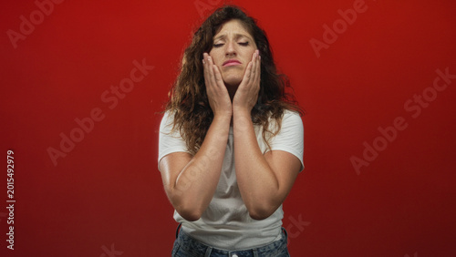 Εκτύπωση καμβά Young hispanic woman seated in white t shirt with pouting mouth in red studio; discomfort hesitation