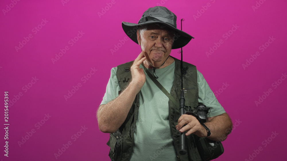 Fototapeta premium Man holding fishing rod with hand on neck for neckache, wearing fishing vest and hat in studio; resilience fatigue.