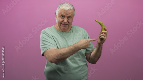 Senior man points finger to banana while holding it with other hand in studio pink backdrop, smiling and engaging directly with camera; playful.