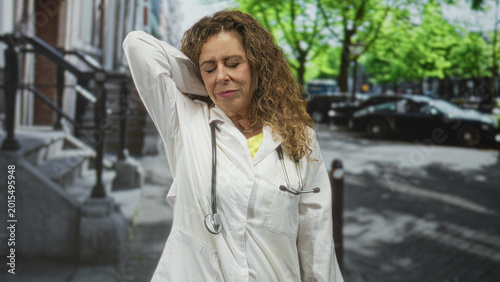 Tableau sur toile Woman doctor in white coat holding neck with hand and stethoscope on city street near brownstone steps and parked cars; fatigue relief