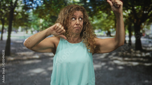 Foto Middleaged woman holding thumbs down and thumbs up with bare forearms on a forest path under trees; indecision