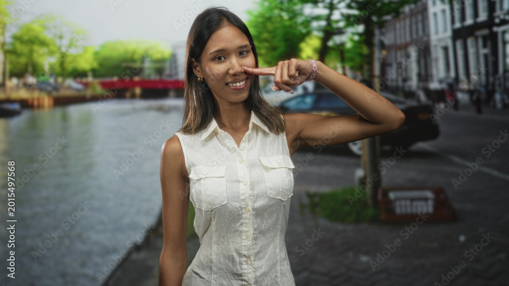 Fototapeta premium Thai young southeast asian brunette woman points finger to cheek on a city street by a canal, wearing white sleeveless blouse and bracelet; playful ease.