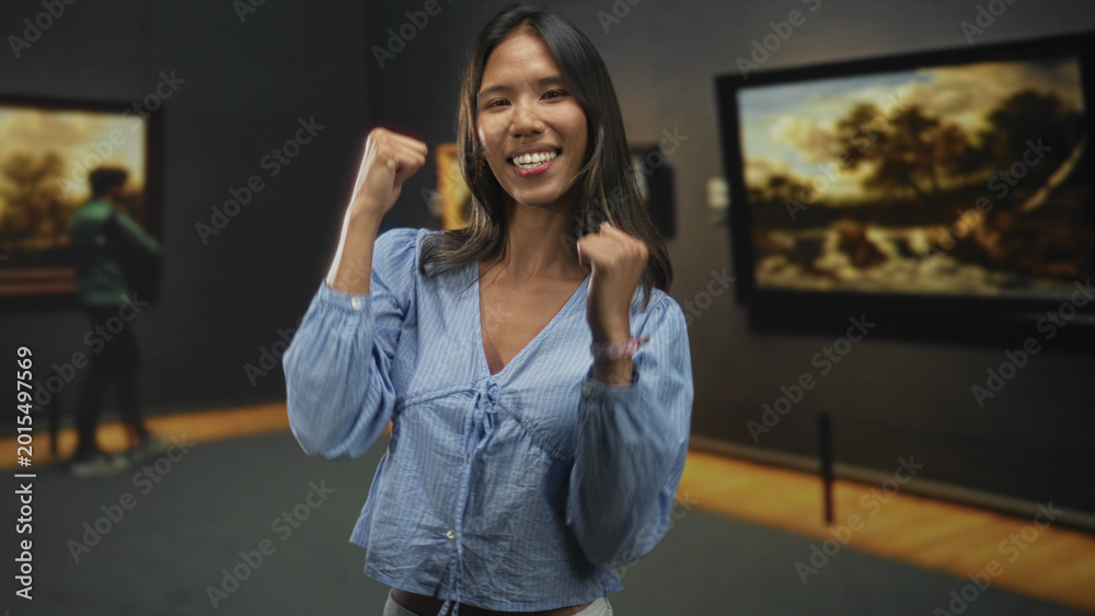 Fototapeta premium Thai woman with fists raised, smiling and dancing in a museum building among paintings; joy celebration.