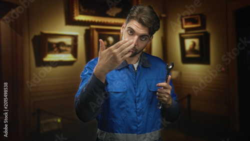 Photography Man mechanic in blue overalls, beard visible, holds wrench and gestures with left hand while inspecting paintings behind a rope stanchion in museum; curiosity duty