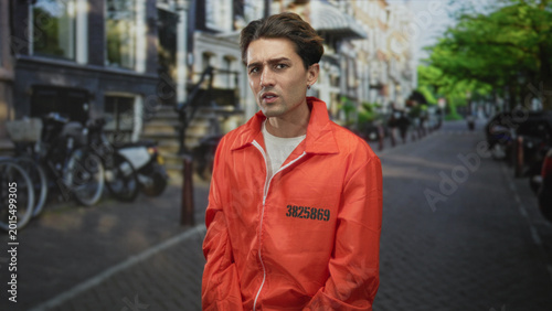 Tablou pe pânză Man in orange prison jumpsuit numbered 3825869 with hands clasped and downcast gaze on a city street; remorse reflection