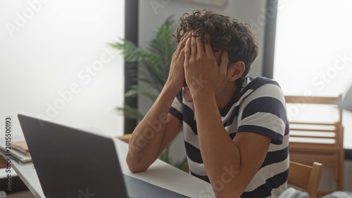 Fotografi Man in striped shirt covering face with hands at desk with laptop and notebooks in building; stress exhaustion