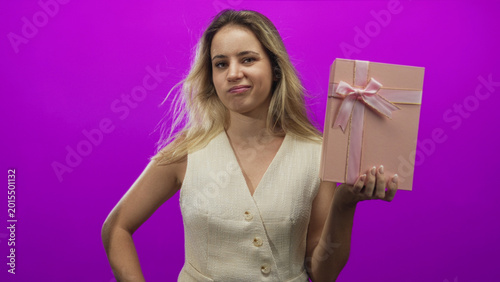 Tableau sur toile Woman holding pink gift box with right hand and hand on hip in magenta studio; annoyance disappointment
