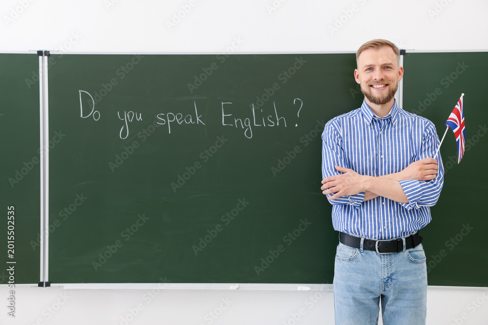 Fototapeta premium English teacher holding flag of United Kingdom at green chalkboard with phrase Do You Speak English? in classroom