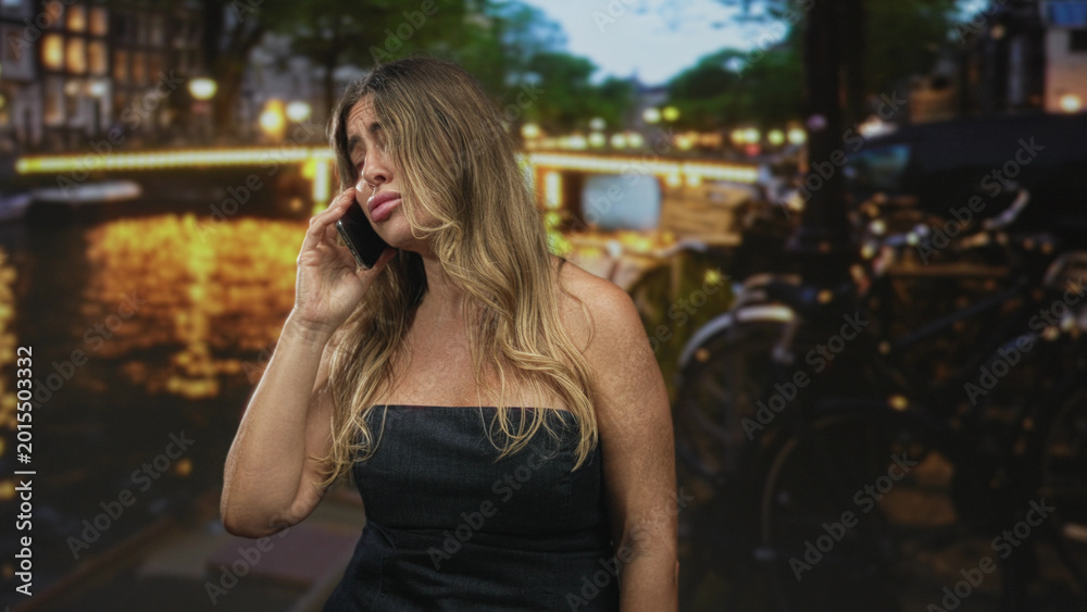 Fototapeta premium Middle aged woman holding phone to ear, eyes closed, wearing strapless black dress on street by lit canal and parked bicycles at night; frustration fatigue.
