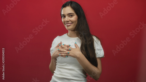 Photography Young hispanic woman in a white shirt places hands on chest in vivid red studio; gratitude warmth sincerity appreciation