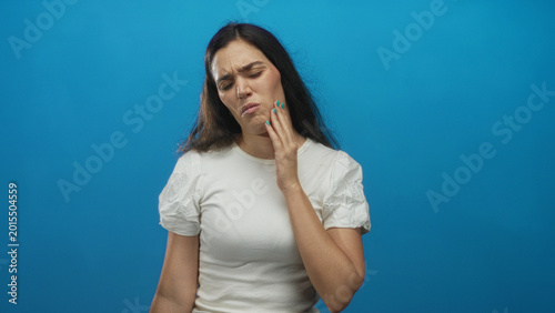 Bild auf Leinwand Young hispanic woman in white tshirt pressing cheek with hand while grimacing in bright blue studio setting; pain