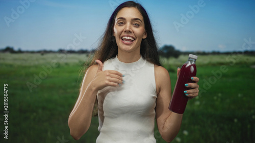 Fotografie Young woman holds a red juice bottle and points finger to bottle in forest; refreshment energy boost vitality