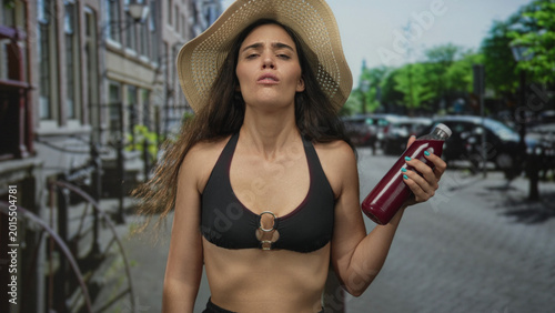 Obraz na plátně Woman in straw hat holds a red juice bottle with crossed arm and pouting lips under bright sunlight on a busy city street; disgust