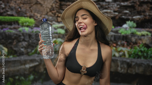 Fotografie Woman in black bikini top and straw hat holds water bottle and sticks out tongue in forest; refreshment