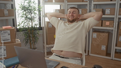 Foto Man at desk surrounded by stacked parcels typing on laptop in building while glancing at phone; small business productivity