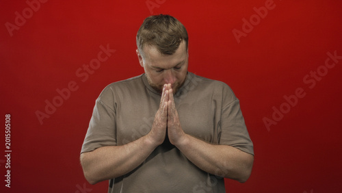 Quadro su tela Young heavyset man with bare hands pressed together in prayer, head bowed, standing against a solid red studio wall; serenity