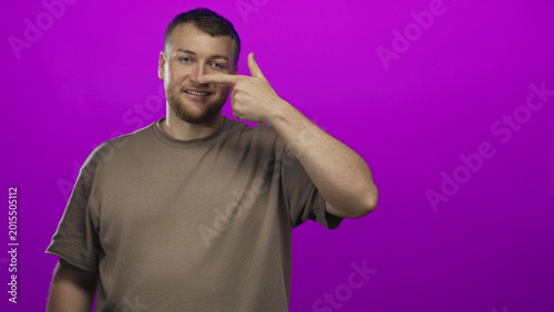 Canvastavla Young caucasian man, heavyset and smiling, wearing brown t shirt, points finger to nose in purple studio; playful