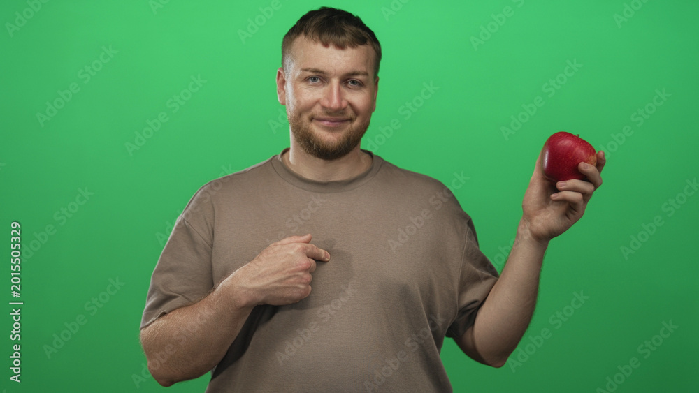 Fototapeta premium Young caucasian man holding a red apple with right hand and thumb pointing to chest in green studio; confidence healthy choice.