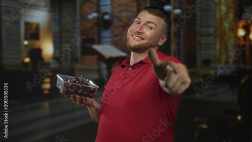 Fotografie Man holding cherries points finger to camera in a church building, smiling and offering fruit; playful invitation