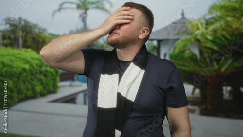 Foto Man holding hand to forehead at resort pool, wearing striped black and white scarf and dark tshirt, facepalm gesture indicating headache; vacation fatigue