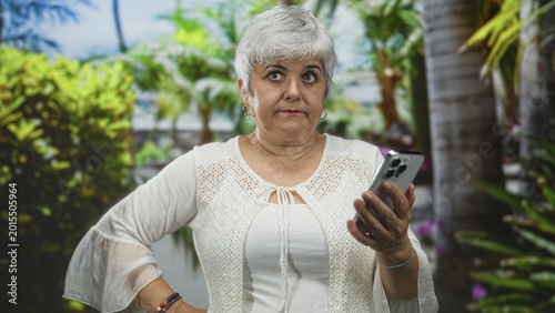 Konstfotografi Woman holding smartphone with hand on hip, frowning while checking phone in forest; technology frustration