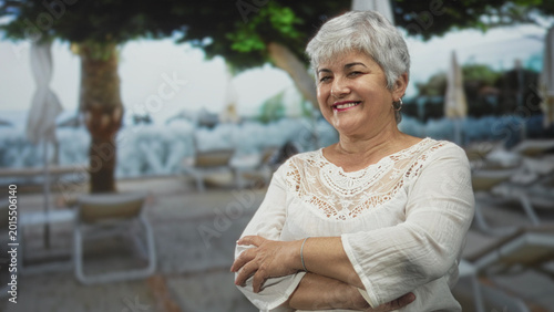 Canvas Print Woman with arms crossed smiling, white lace top and bracelet on a seaside street with loungers and umbrellas; contentment relaxation
