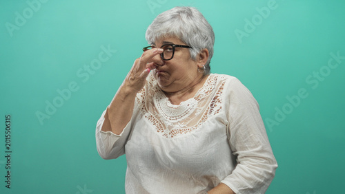 Fotografija Woman senior with grey hair wearing spectacles and white blouse pinching nose with hand in studio against teal backdrop; disgust reaction