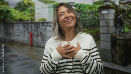 Cuadro en lienzo Middle age hispanic woman clasping hands over chest on a wet residential street, smiling with closed eyes; gratitude warmth relief