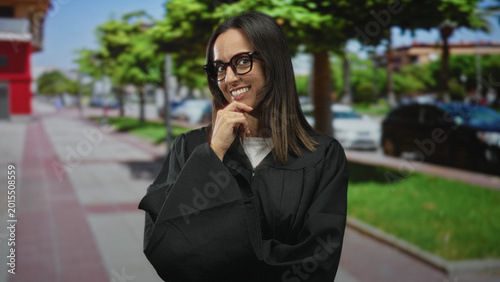 Fotografía Woman judge in black robe wearing glasses with hand on chin smiling on a sunny urban street; confidence thoughtfulness professional