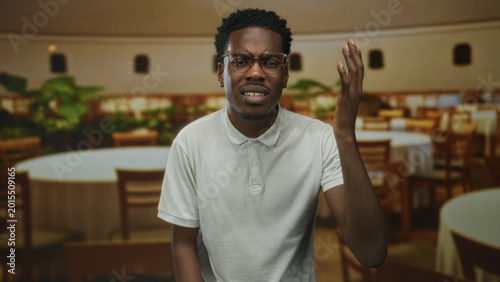 Foto African american man wearing white polo shirt raising right hand gesture inside restaurant building; confusion