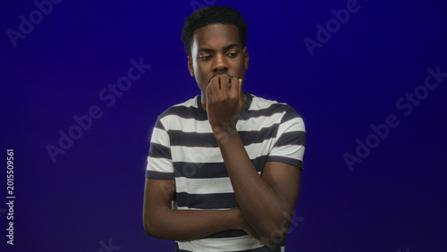 Bild auf Leinwand Young african american man biting fingernails with hand to mouth and tense posture in blue studio space; anxiety