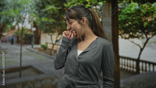 Photos Young woman biting her hand and smiling, fingernails visible, wearing gray cardigan, standing on a street with trees and stone balustrade; playful shyness