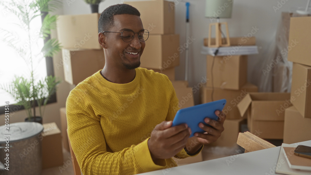 Αυτοκόλλητο Young black man with glasses wearing yellow sweater holding and tapping a blue tablet while seated at a table surrounded by packed cardboard moving boxes in apartment; contentment settling